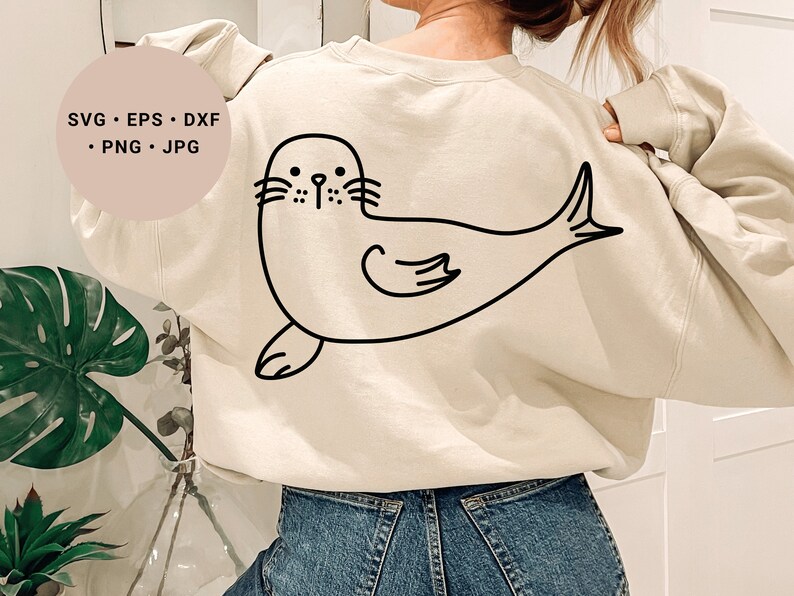 May include: A cream-colored sweatshirt with a black line drawing of a seal on the back. The seal is facing the left side of the image and has a simple, cartoon-like design.