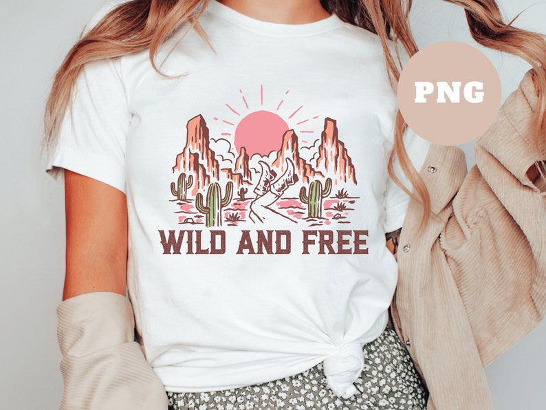 Puede incluir: Camiseta blanca con un dise&ntilde;o gr&aacute;fico de un paisaje des&eacute;rtico con un sol rosa, monta&ntilde;as y cactus. El texto "WILD AND FREE" est&aacute; impreso debajo del gr&aacute;fico.