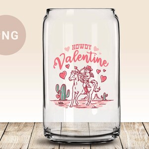 May include: A clear glass can with a pink and red design featuring a cowboy cupid riding a horse with the text "Howdy Valentine".
