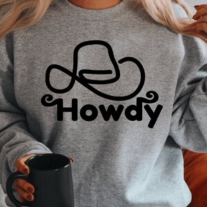 Może przedstawiać: Szara bluza z czarnym konturem kowbojskiego kapelusza i nadrukiem słowa "Howdy" z przodu.