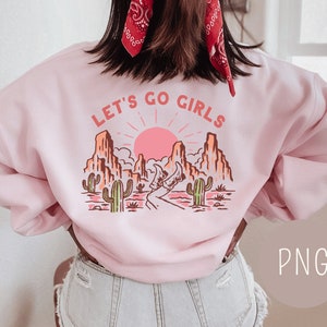 May include: Pink sweatshirt with a graphic of a desert landscape and the text "Let's Go Girls".