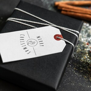 May include: A white gift tag with black text that reads "Merry Christmas" and a simple illustration of a Christmas tree. The tag is tied to a black gift box with white twine.