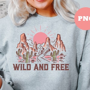 Puede incluir: Una sudadera gris con un dise&ntilde;o gr&aacute;fico de un paisaje des&eacute;rtico con un sol rosa, cactus y monta&ntilde;as. El texto "WILD AND FREE" est&aacute; impreso debajo del gr&aacute;fico.