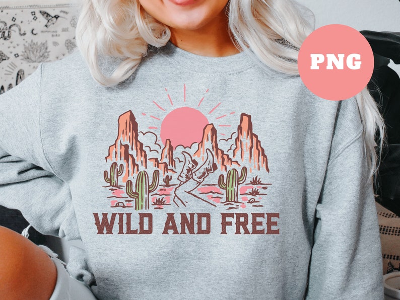 Puede incluir: Una sudadera gris con un dise&ntilde;o gr&aacute;fico de un paisaje des&eacute;rtico con un sol rosa, cactus y monta&ntilde;as. El texto "WILD AND FREE" est&aacute; impreso debajo del gr&aacute;fico.