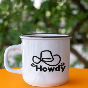 Może przedstawiać: Biały kubek ceramiczny z czarną obw&oacute;dką i czarnym wzorem w postaci kowbojskiego kapelusza z napisem "Howdy" pod spodem.