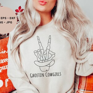 May include: A white sweatshirt with a black line drawing of a skeleton hand making a peace sign. The text "Ghostin Cowgirls" is printed below the hand.