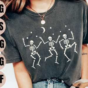 May include: A dark grey t-shirt featuring a white graphic of three skeletons dancing beneath a crescent moon and stars. The skeletons are depicted in a simple design without facial features.