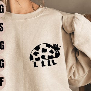 Könnte beinhalten: Ein schwarz-weißes Kuhmuster-Design einer Marienkäferin, die einen Cowboyhut trägt. Der Marienkäfer hat vier Beine und trägt Cowboystiefel. Das Design befindet sich auf einem hellbeigen Sweatshirt.