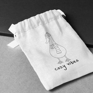 K&ouml;nnte beinhalten: Ein wei&szlig;er Kordelzugbeutel mit einer schwarzen Strichzeichnung einer Gans, die einen Hut und einen Schal tr&auml;gt. Der Text "cozy vibes" ist unter der Gans aufgedruckt.
