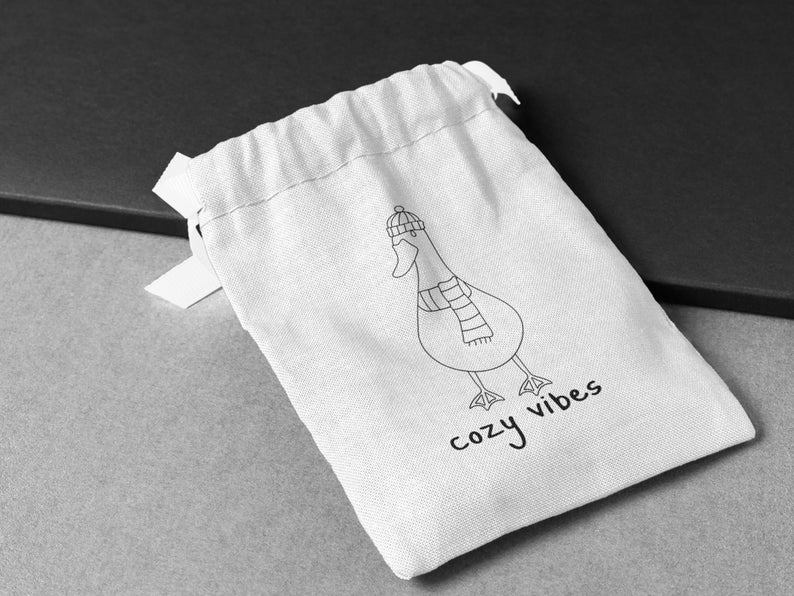 K&ouml;nnte beinhalten: Ein wei&szlig;er Kordelzugbeutel mit einer schwarzen Strichzeichnung einer Gans, die einen Hut und einen Schal tr&auml;gt. Der Text "cozy vibes" ist unter der Gans aufgedruckt.