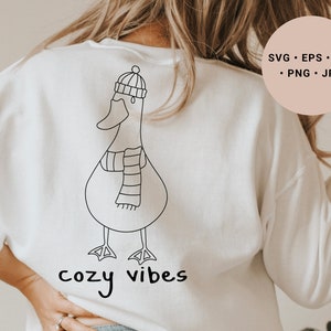 K&ouml;nnte beinhalten: Wei&szlig;er Pullover mit einer schwarzen Linienzeichnung eines Entchens mit Hut und Schal. Der Text "cozy vibes" ist unter dem Entchen gedruckt.