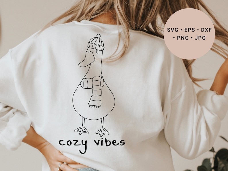 K&ouml;nnte beinhalten: Wei&szlig;er Pullover mit einer schwarzen Linienzeichnung eines Entchens mit Hut und Schal. Der Text "cozy vibes" ist unter dem Entchen gedruckt.
