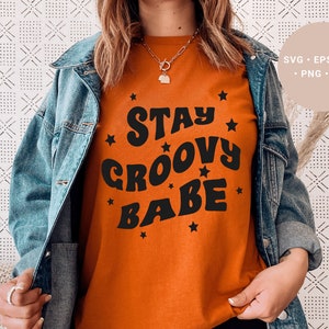 May include: A woman wearing a burnt orange t-shirt with the text "Stay Groovy Babe" in black retro font and surrounded by small black stars. She is wearing a blue denim jacket.
