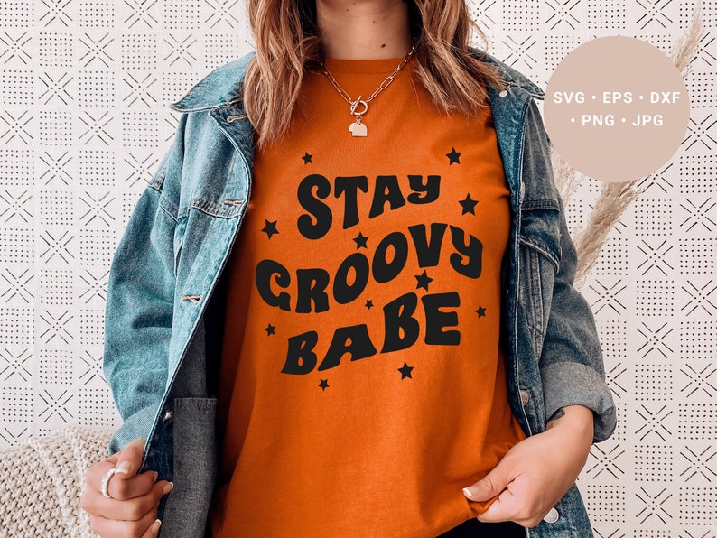 May include: A woman wearing a burnt orange t-shirt with the text "Stay Groovy Babe" in black retro font and surrounded by small black stars. She is wearing a blue denim jacket.