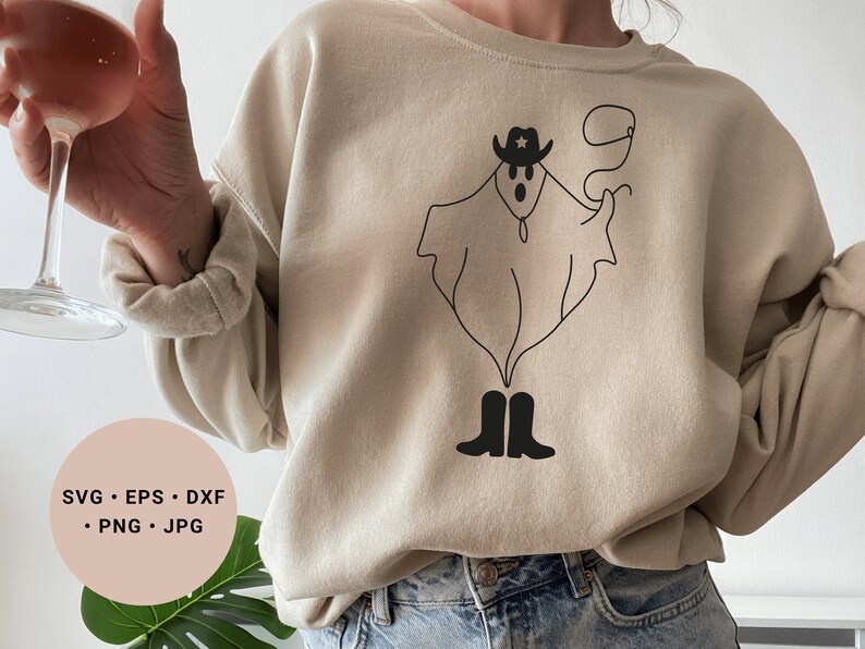 May include: A beige sweatshirt with a black line drawing of a ghost wearing a cowboy hat and boots. The ghost is holding a lasso. The text "SVG - EPS - DXF - PNG - JPG" is on a pink circle in the lower left corner of the image.