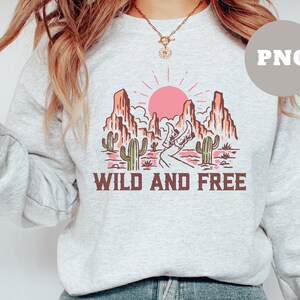 Puede incluir: Una sudadera gris con un dise&ntilde;o gr&aacute;fico de un paisaje des&eacute;rtico con un sol rosa, cactus y monta&ntilde;as. El texto "WILD AND FREE" est&aacute; impreso debajo del gr&aacute;fico.