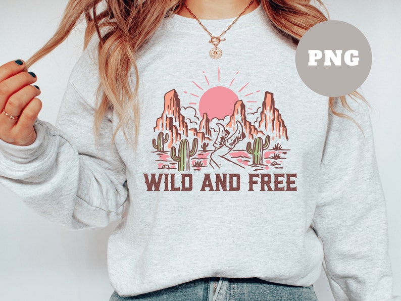 Puede incluir: Una sudadera gris con un dise&ntilde;o gr&aacute;fico de un paisaje des&eacute;rtico con un sol rosa, cactus y monta&ntilde;as. El texto "WILD AND FREE" est&aacute; impreso debajo del gr&aacute;fico.