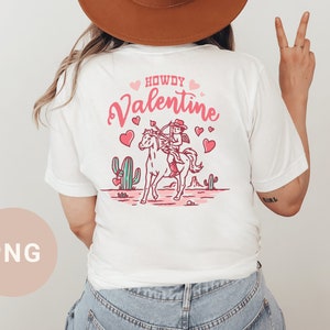 May include: White t-shirt with a pink and red graphic of a cowboy riding a horse with the text "Howdy Valentine".