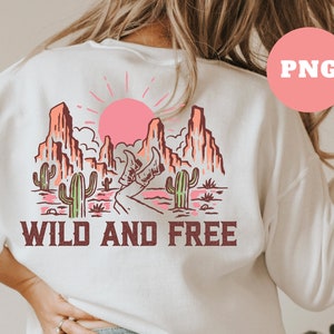 Puede incluir: Sudadera blanca con un estampado gr&aacute;fico de un paisaje des&eacute;rtico con monta&ntilde;as, un sol poniente, cactus y el texto "WILD AND FREE".