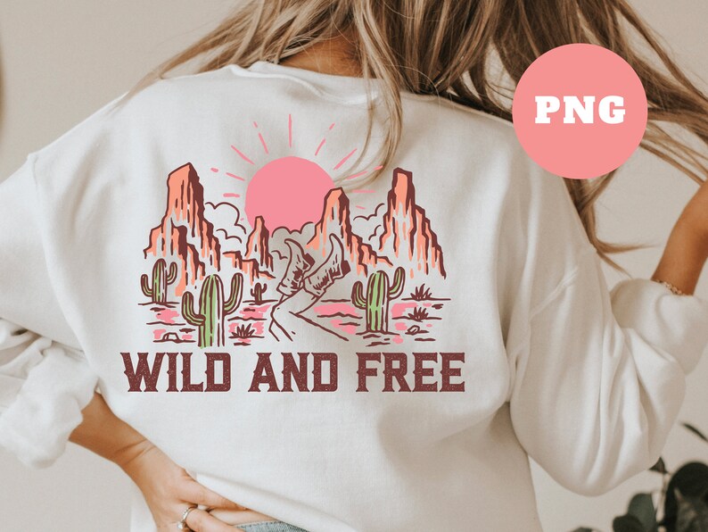 Puede incluir: Sudadera blanca con un estampado gr&aacute;fico de un paisaje des&eacute;rtico con monta&ntilde;as, un sol poniente, cactus y el texto "WILD AND FREE".