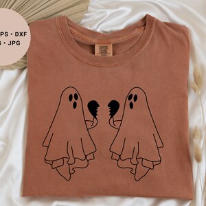May include: A brown t-shirt with a black graphic of two ghosts holding broken heart halves. The ghosts are facing each other and have three dots for eyes.