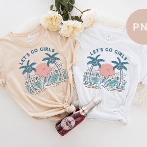 K&ouml;nnte beinhalten: Zwei T-Shirts, eines beige und eines wei&szlig;, jeweils mit einer Strandmotiv-Grafik. Das Design umfasst Palmen, einen Sonnenuntergang und den Text "LET'S GO GIRLS" in kreisf&ouml;rmiger Anordnung. Eine Flasche Sekt befindet sich im Vordergrund.