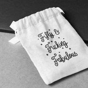 May include: A white drawstring gift bag with the text "Fifty & Fucking Fabulous" in black script. The bag has a simple design with a small ribbon tie and is set against a gray and black background.