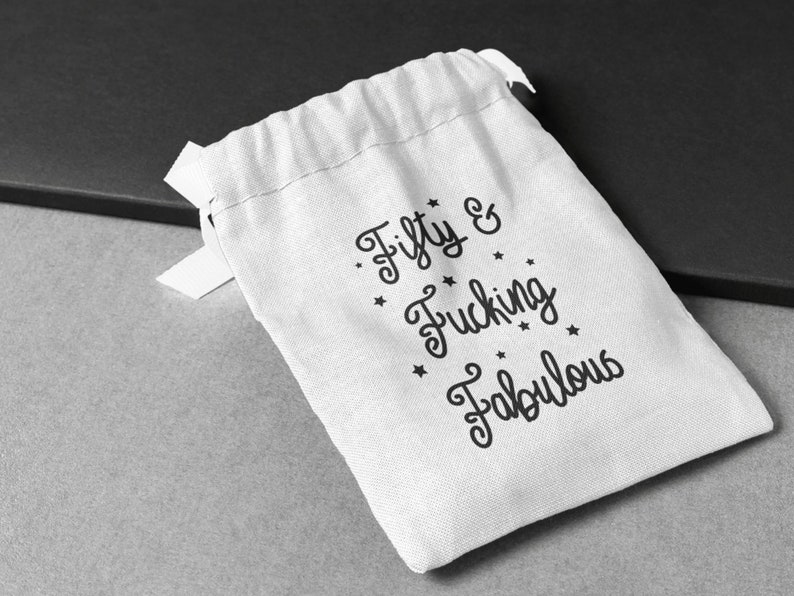 May include: A white drawstring gift bag with the text "Fifty & Fucking Fabulous" in black script. The bag has a simple design with a small ribbon tie and is set against a gray and black background.