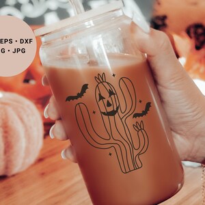 May include: A glass jar containing a brown drink. The jar features a black and white illustration of a cactus with a jack-o'-lantern face and bats flying around it. The jar is being held by a hand wearing a bracelet with a ghost charm.