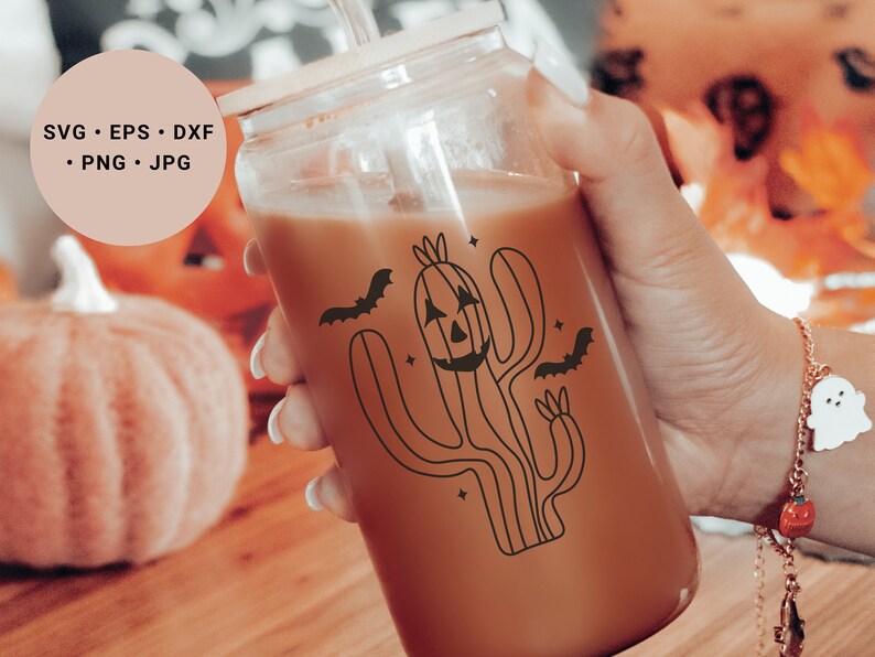 May include: A glass jar containing a brown drink. The jar features a black and white illustration of a cactus with a jack-o'-lantern face and bats flying around it. The jar is being held by a hand wearing a bracelet with a ghost charm.