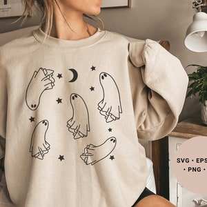 May include: A beige sweatshirt featuring a graphic design of five ghosts and stars. The ghosts are outlined in black and have a simple, whimsical design. The text "SVG - EPS - DXF - PNG - JPG" is printed on a pink background in the bottom right corner of the image.