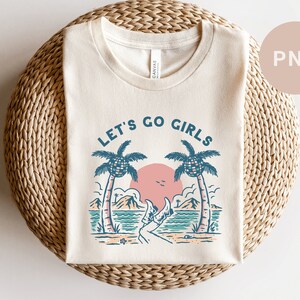 K&ouml;nnte beinhalten: Cremefarbenes T-Shirt mit Strandmotiv. Das Design zeigt Palmen, eine rosa Sonne und den Text "LET'S GO GIRLS" in Blau. Das Shirt liegt gefaltet auf einer gewebten, kreisf&ouml;rmigen Oberfl&auml;che.