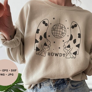 Könnte beinhalten: Ein beiger Pullover mit einem schwarzen und weißen Kuhflecken-Geisterdesign, einer Discokugel und dem Wort "HOWDY" auf der Vorderseite.