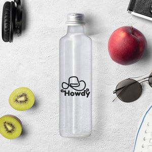 Może przedstawiać: Przezroczysta butelka szklana z srebrną zakrętką i czarno-białym logo z napisem "Howdy" i wzorem kowbojskiego kapelusza. Butelka stoi na białym tle z czerwonym jabłkiem, kiwi, okularami przeciwsłonecznymi, słuchawkami i aparatem.