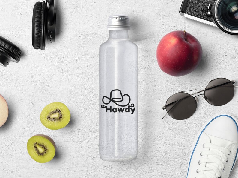 Może przedstawiać: Przezroczysta butelka szklana z srebrną zakrętką i czarno-białym logo z napisem "Howdy" i wzorem kowbojskiego kapelusza. Butelka stoi na białym tle z czerwonym jabłkiem, kiwi, okularami przeciwsłonecznymi, słuchawkami i aparatem.