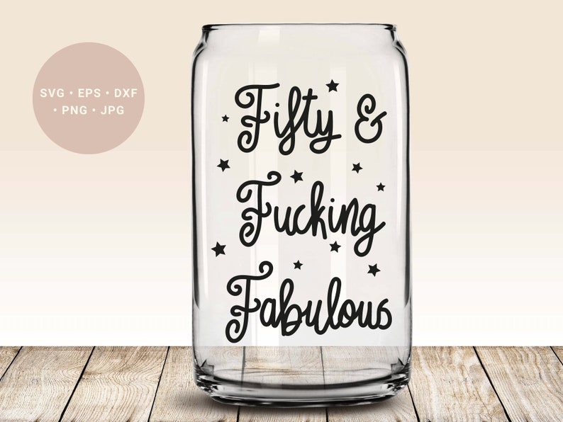 May include: A clear glass can-shaped drinking glass with the words "Fifty & Fucking Fabulous" in black script, surrounded by small black star shapes. The glass sits on a wooden surface.