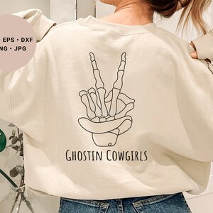 May include: A cream-colored sweatshirt with a black line drawing of a skeleton hand making a peace sign. The text "Ghostin Cowgirls" is printed below the hand.