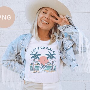 K&ouml;nnte beinhalten: Wei&szlig;es T-Shirt mit dem Text "LET'S GO GIRLS" und einer Strandgrafik. Die Grafik zeigt Palmen, einen Sonnenuntergang und eine Person. Die Person tr&auml;gt einen Hut und eine Jeansjacke.
