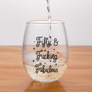 May include: A clear stemless wine glass with the words "Fifty & Fucking Fabulous" in black script. The glass is being filled with a clear liquid, likely wine, and is set on a wooden surface.