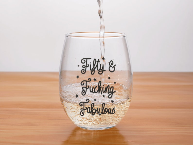 May include: A clear stemless wine glass with the words "Fifty & Fucking Fabulous" in black script. The glass is being filled with a clear liquid, likely wine, and is set on a wooden surface.