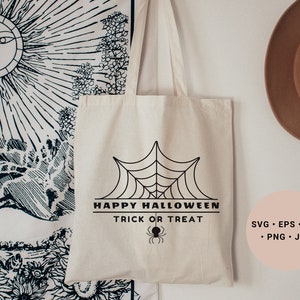 K&ouml;nnte beinhalten: Eine wei&szlig;e Stofftasche mit einem schwarzen Spinnennetz-Design und dem Text "Happy Halloween Trick or Treat". Die Tasche h&auml;ngt an einer wei&szlig;en Wand mit einer Wandbehang und einem braunen Hut im Hintergrund.