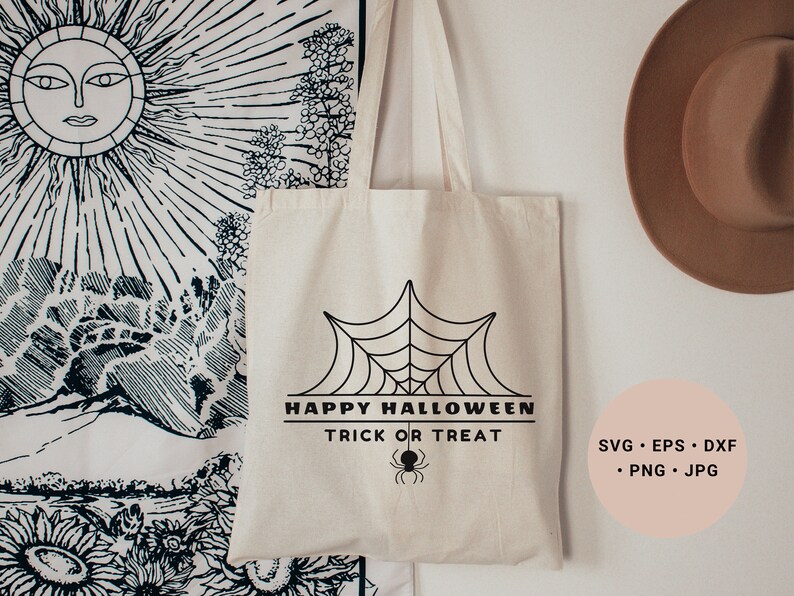 K&ouml;nnte beinhalten: Eine wei&szlig;e Stofftasche mit einem schwarzen Spinnennetz-Design und dem Text "Happy Halloween Trick or Treat". Die Tasche h&auml;ngt an einer wei&szlig;en Wand mit einer Wandbehang und einem braunen Hut im Hintergrund.