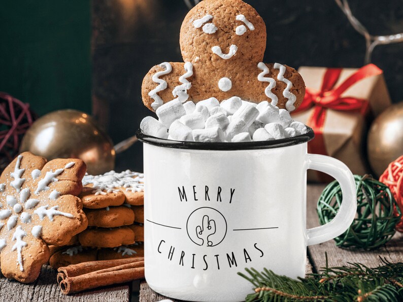 May include: White enamel mug with black trim and the text "Merry Christmas" with a stylised cactus design. The mug is filled with marshmallows and topped with a gingerbread man cookie.