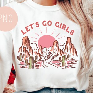 May include: White sweatshirt with a pink, orange, and brown graphic design of a desert landscape with a rising sun and the text "LET'S GO GIRLS".