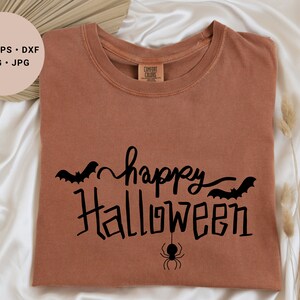 Puede incluir: Una camiseta marrón con el texto "happy halloween" en letra negra con murciélagos y un gráfico de araña.