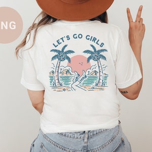 K&ouml;nnte beinhalten: Wei&szlig;es T-Shirt mit Strandmotiv. Das Design zeigt Palmen, einen Sonnenuntergang und den Text "LET'S GO GIRLS". Das Shirt ist ein l&auml;ssiger Stil, geeignet f&uuml;r den Alltag.
