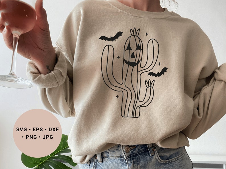 May include: A beige sweatshirt featuring a black outline of a cactus with a jack-o'-lantern face and bats flying around it. The text "SVG - EPS - DXF - PNG - JPG" is on a pink circle in the bottom left corner of the image.