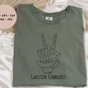 May include: A green t-shirt with a black line drawing of a skeleton hand making a peace sign. The skeleton hand is wearing a cowboy hat. The text "Ghostin Cowgirls" is printed below the hand.