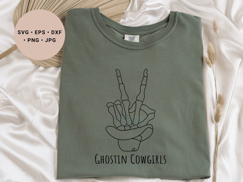 May include: A green t-shirt with a black line drawing of a skeleton hand making a peace sign. The skeleton hand is wearing a cowboy hat. The text "Ghostin Cowgirls" is printed below the hand.