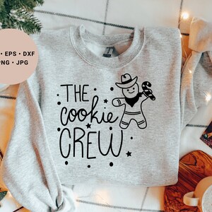 May include: A gray sweatshirt with the text "THE cookie CREW" in black lettering. A gingerbread man wearing a cowboy hat and holding a candy cane is above the text.